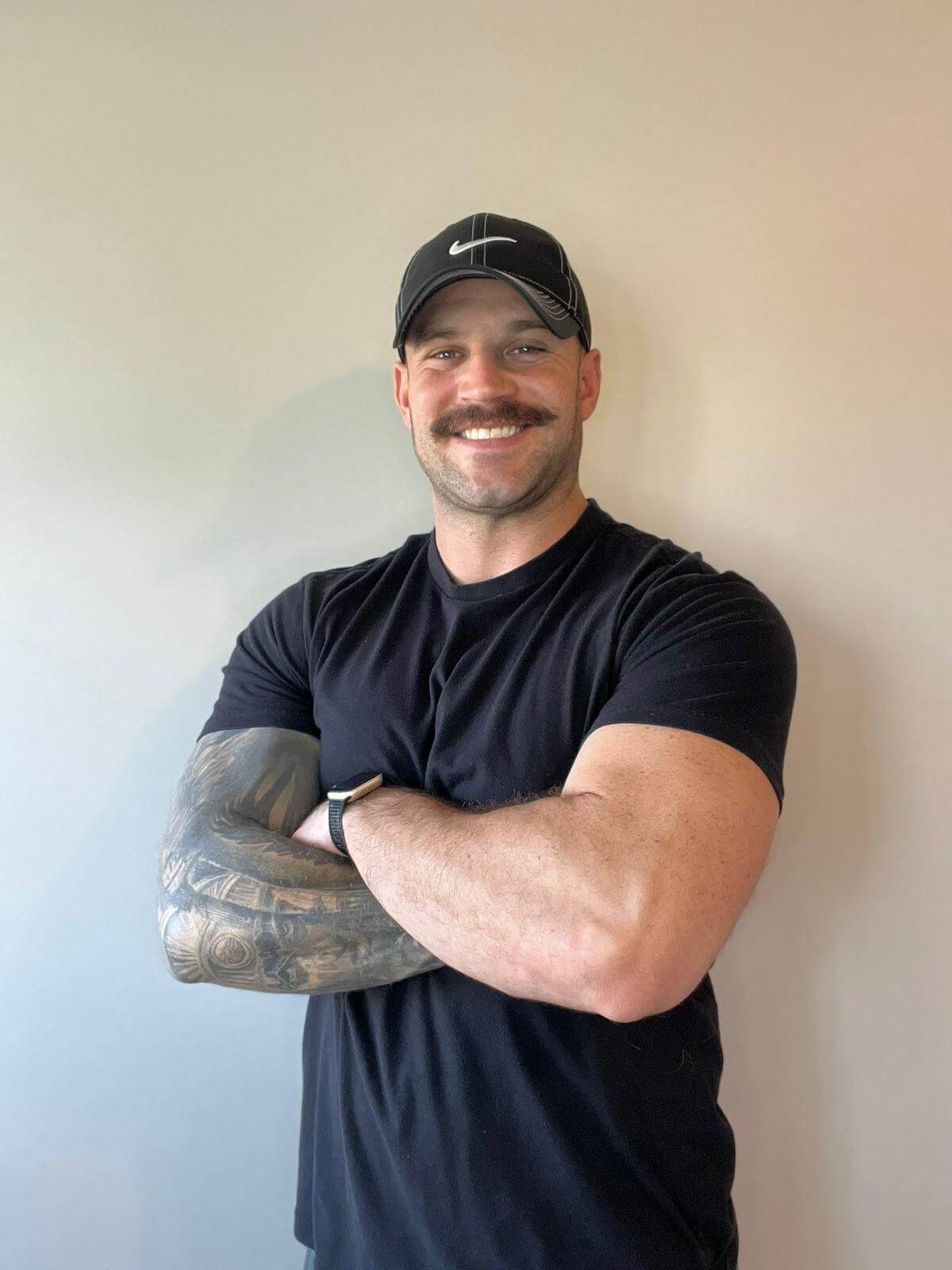 Coach Mario with his arms crossed in front of a white wall, wearing a plain black t shirt and black baseball cap